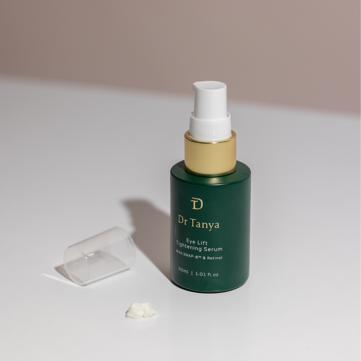 An emerald bottle of eye lift serum with the clear cap lying next to it and a drop of serum in front of it