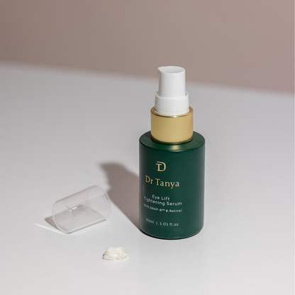 An emerald bottle of eye lift serum with the clear cap lying next to it and a drop of serum in front of it