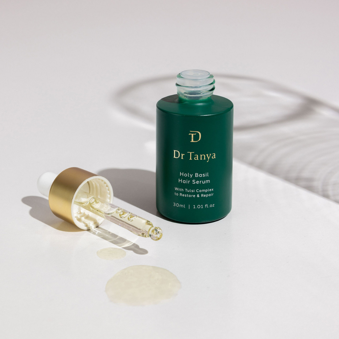 An emerald green bottle of hair serum with the dropper cap lying next to it