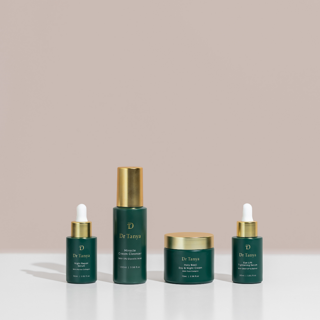 Four dark green skincare product bottles with gold lids lined up in a row against a soft pink backdrop