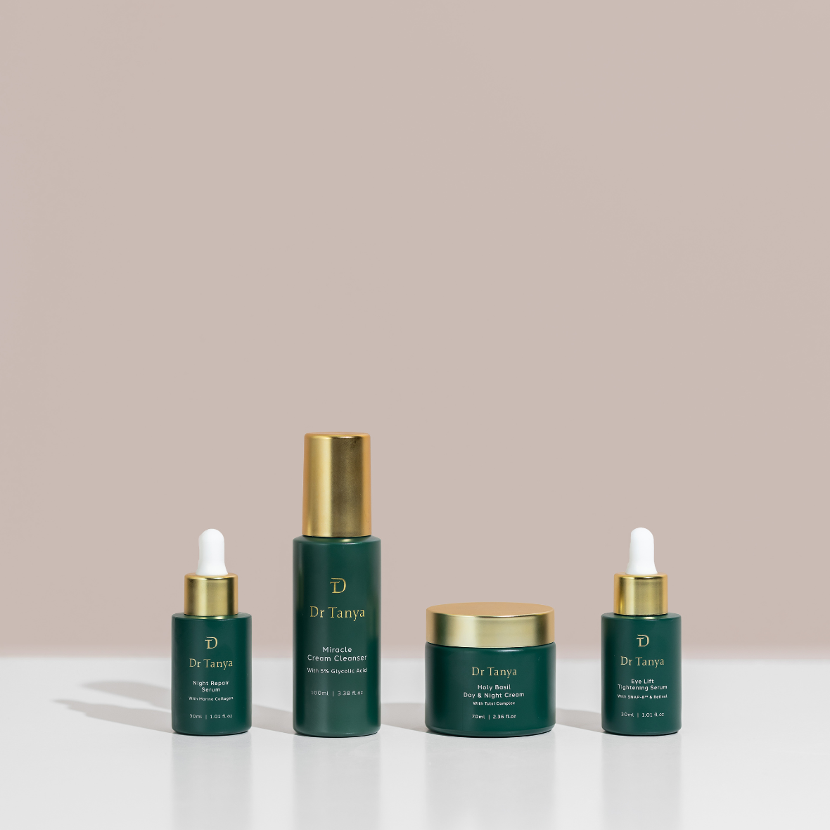 Four dark green skincare product bottles with gold lids lined up in a row against a soft pink backdrop